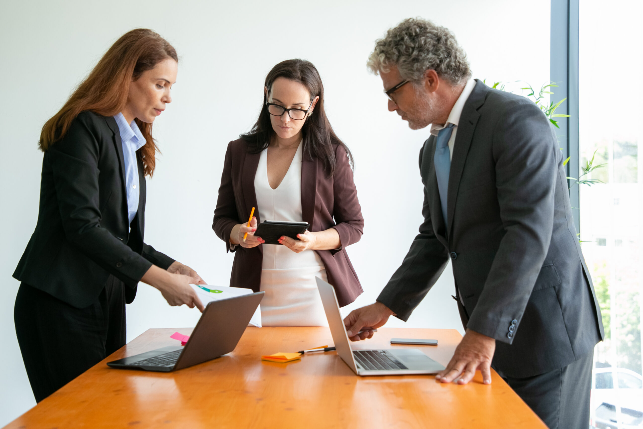 Senior manager in eyeglasses discussing with businesswomen and using laptop. Successful content businesspeople standing and talking together in conference room. Business and collaboration concept